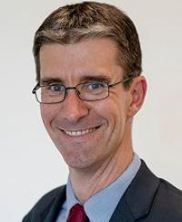 Simon Duggan. He is smiling wearing glasses and a dark suit with red tie.