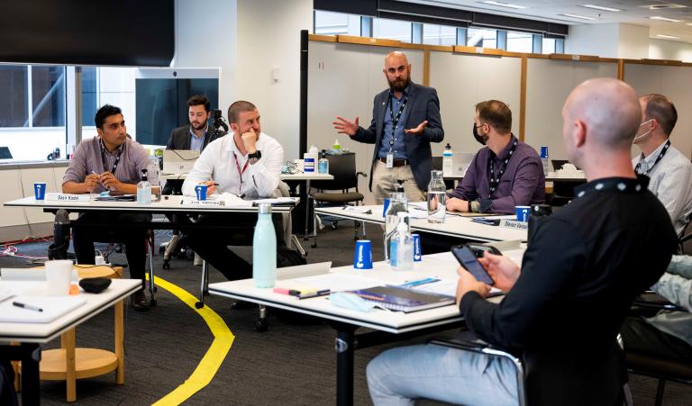 Room of six participants with a Facilitator. All bar one are seated around a circle of desks with a whiteboard in background of room.