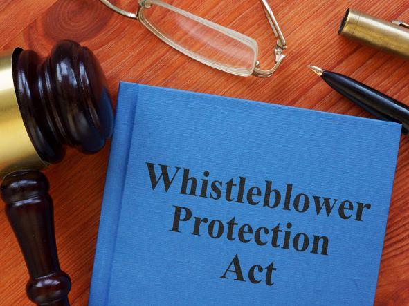 Image of hard cover blue book saying, “Whistleblower Protection Act”, with a gavel next to it on desktop with pen, reading glasses.