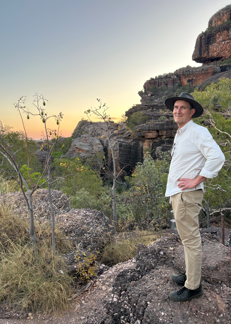 A photo of a male standing on a rocky outcrop with Australian rockscape behind him.