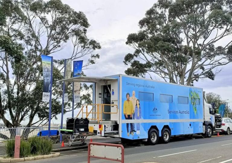 Image of Services Australia bus parked by side of road with picture of three people painted on its side with blue gum tree leaves on the background.