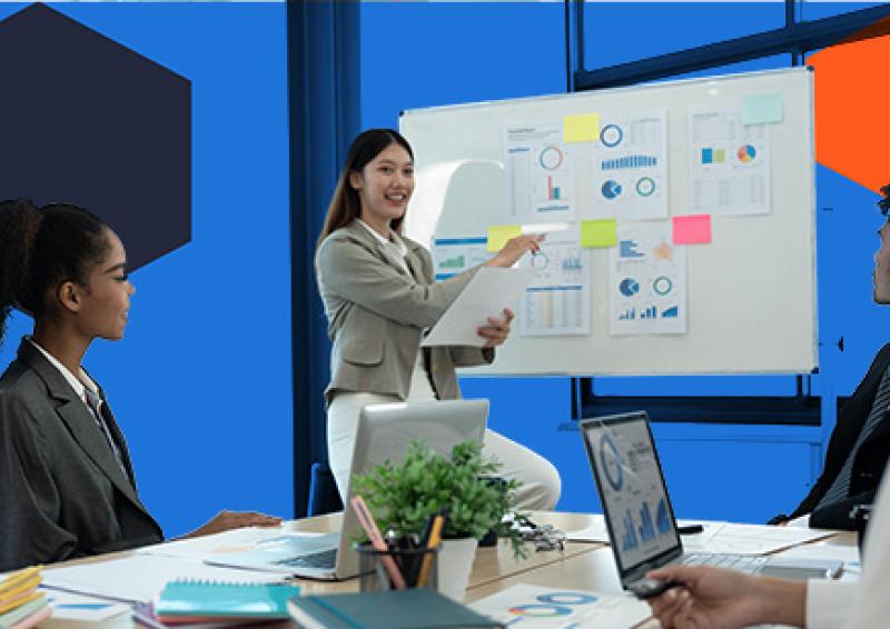 Three people sitting around a desk with a person pointing to graphs on a whiteboard.
