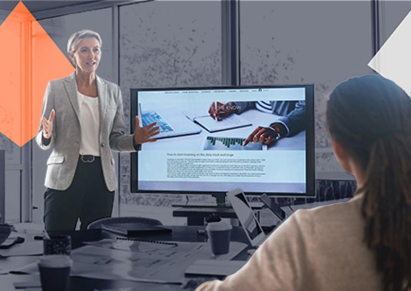 Person presenting in front of a screen with two people sitting at a meeting desk.