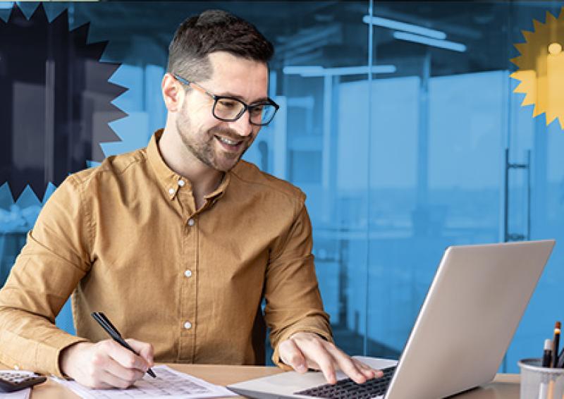 Person sitting at a desk working on a laptop and writing on a piece of paper