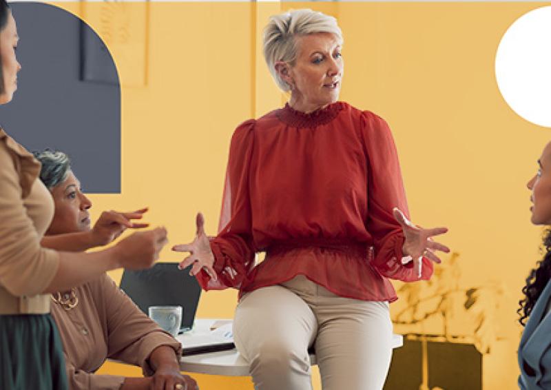 Person sitting on the edge of a desk, hands in an open palm gesture talking to three people.