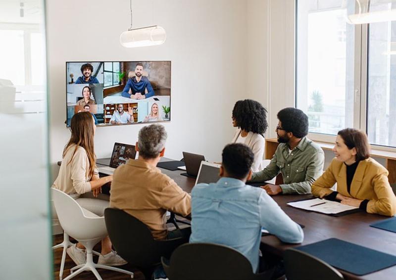 Image of a hybrid event. Six people sitting in a meeting room looking at a wall mounted screen showing other people participating in the event remotely.