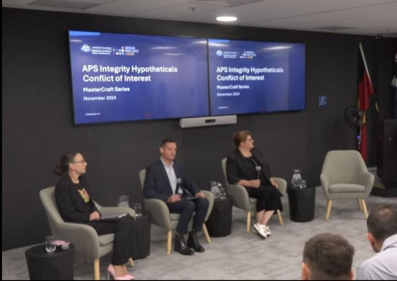 3 panel members sitting on a stage under TV's displaying "APS Integrity Hypotheticals Conflict of Interest. MasterCraft Series"