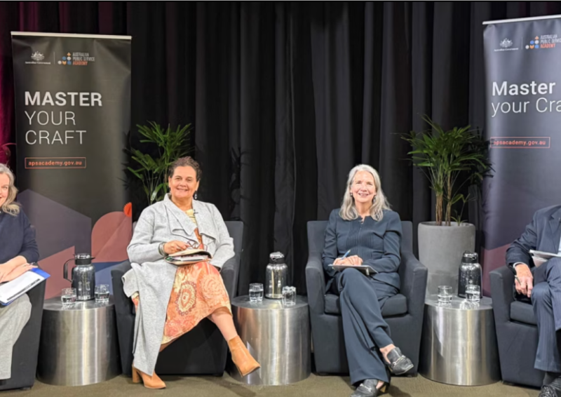Photo of 4 panel members seated together in bucket chairs on stage facing the camera inside an auditorium.