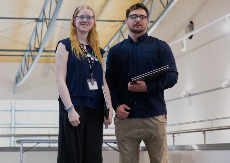 An image of 2 APS Academy Campuses graduates – woman and man - standing facing the camera inside a large auditorium.