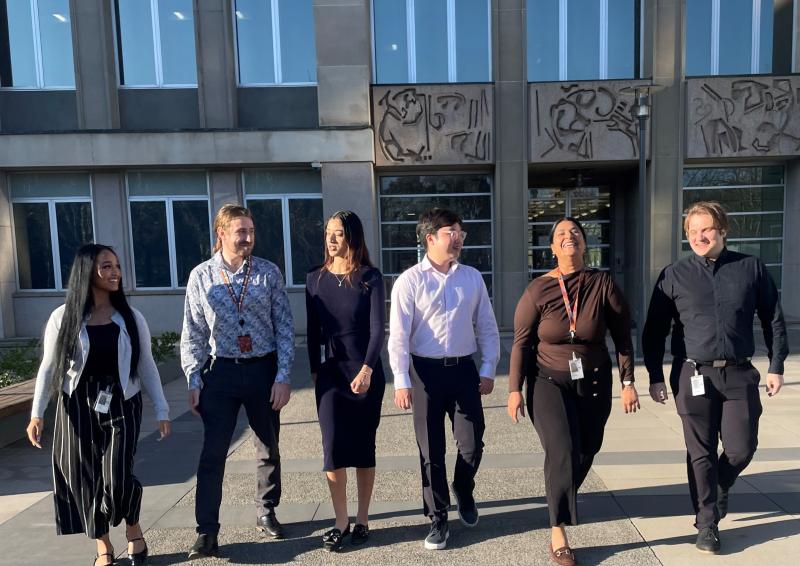 A team of six smiling and chatting APS staff of different genders, ethnicities and racial features leave their APS workplace together with a happy demeanour.