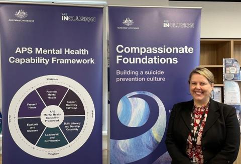 Rachael McMahon stands beside two banners. One says APS Mental Health Capability Framework and the other says Compassionate Foundations: Building a suicide prevention culture.