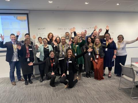 a group of 19 people inside an office with hands in the air smiling for the camera.