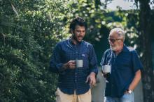 Two men holding coffee cups, walking along next to some tree foliage.