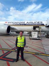A man, Eamon Lindsay, in high visibility vest standing in front of a plane having cargo loaded.