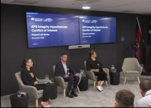 3 panel members sitting on a stage under TV's displaying "APS Integrity Hypotheticals Conflict of Interest. MasterCraft Series"