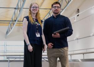 An image of 2 APS Academy Campuses graduates – woman and man - standing facing the camera inside a large auditorium. 