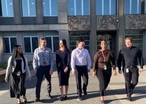 A team of six smiling and chatting APS staff of different genders, ethnicities and racial features leave their APS workplace together with a happy demeanour.