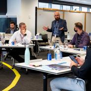 Room of six participants with a Facilitator. All bar one are seated around a circle of desks with a whiteboard in background of room.