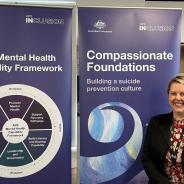 Rachael McMahon stands beside two banners. One says APS Mental Health Capability Framework and the other says Compassionate Foundations: Building a suicide prevention culture.