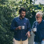 Two men holding coffee cups, walking along next to some tree foliage.