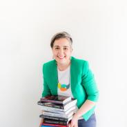 Portrait photo of Clare Conroy smiling while holding a large pile of books