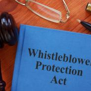 Image of hard cover blue book saying, “Whistleblower Protection Act”, with a gavel next to it on desktop with pen, reading glasses.
