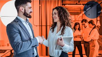 Two people standing shaking hands in the foreground.  Three people standing looking at a tablet.