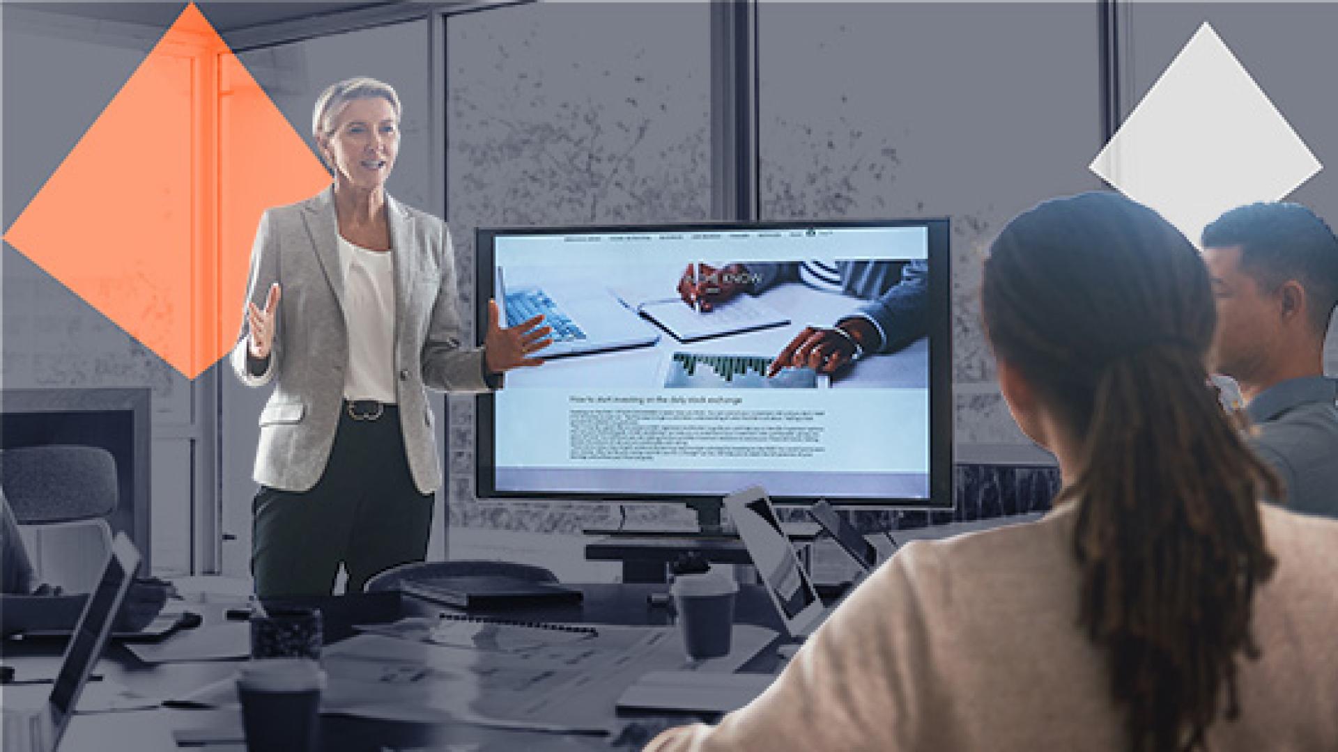 Person presenting in front of a screen with two people sitting at a meeting desk.