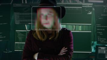 Photo of a person with face pixelated standing in a darkened room in front of a bank of monitors and computer equipment.