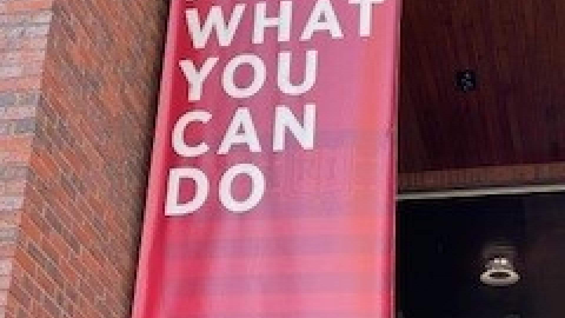 Photo of hanging wall sign affixed to a brick wall saying, ‘Ask what you can do’.