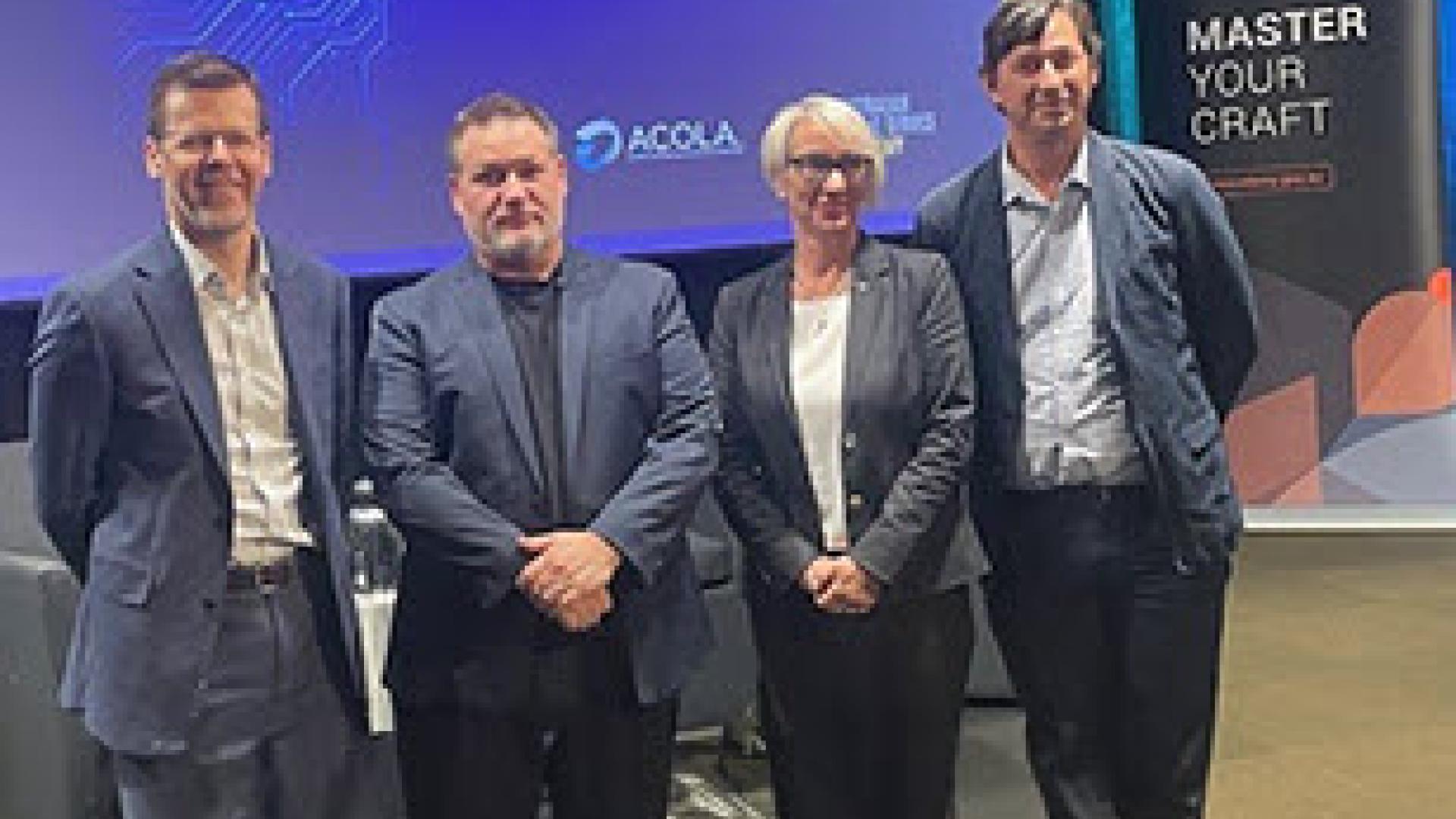Photo taken of four speakers inside the Department of Education seminar room with stage and organisational banners behind them. 