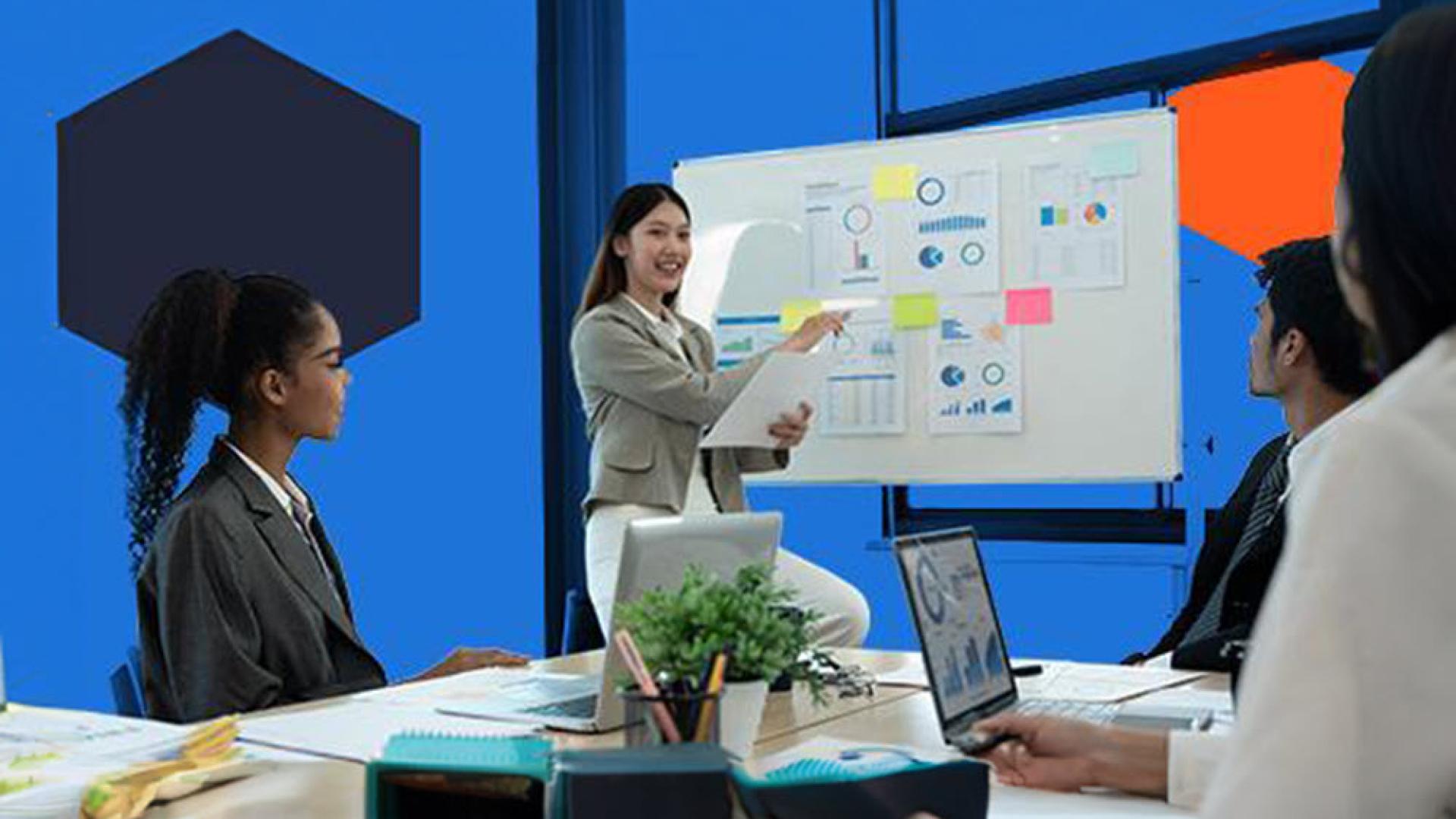 Three people sitting around a desk with a person pointing to graphs on a whiteboard.
