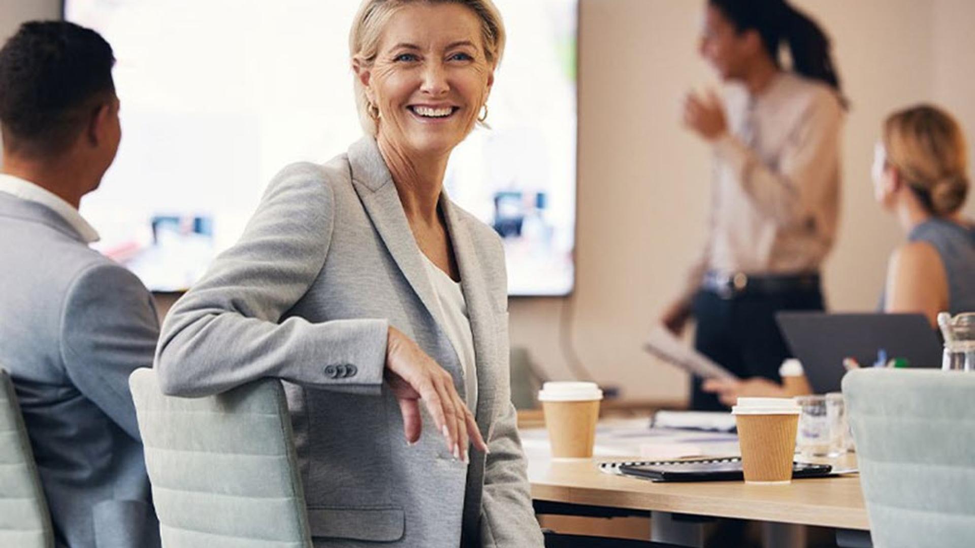 Photo of people sitting around a meeting desk look at a presentation, with a person smiling and looking directly in the camera
