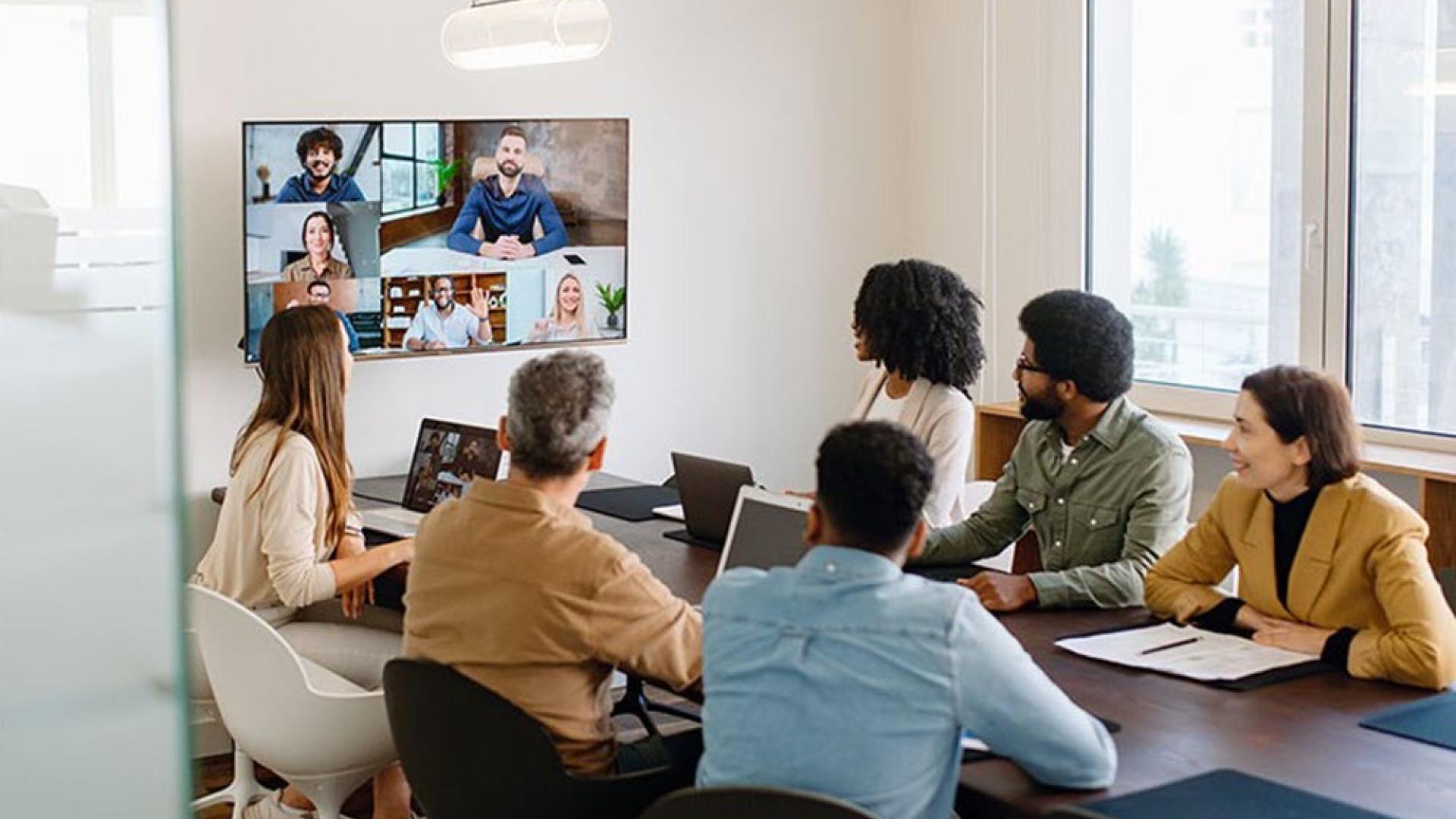 Image of a hybrid event. Six people sitting in a meeting room looking at a wall mounted screen showing other people participating in the event remotely.