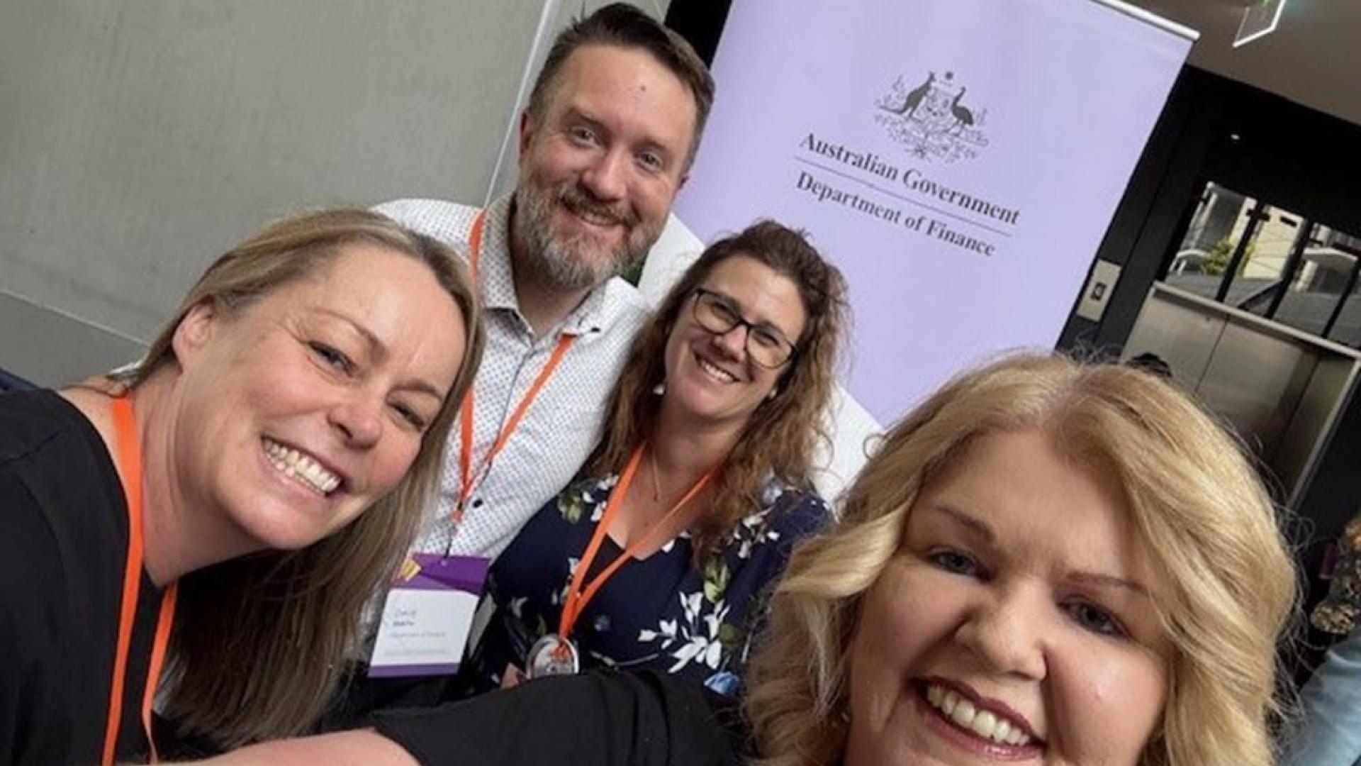 Photo of 4 people taken in front of a Department of Finance pull up banner with logo on it.