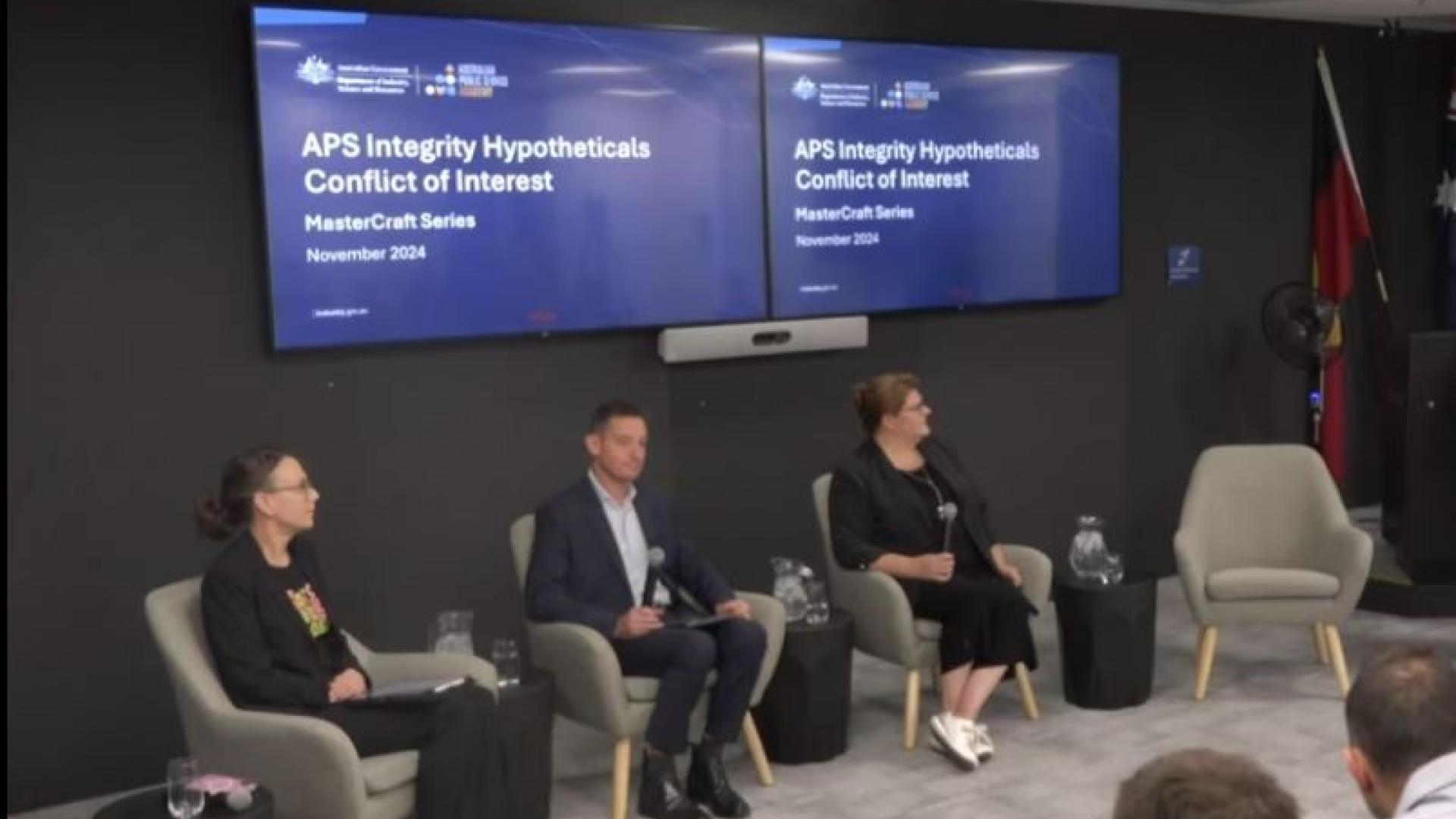 3 panel members sitting on a stage under TV's displaying "APS Integrity Hypotheticals Conflict of Interest. MasterCraft Series"