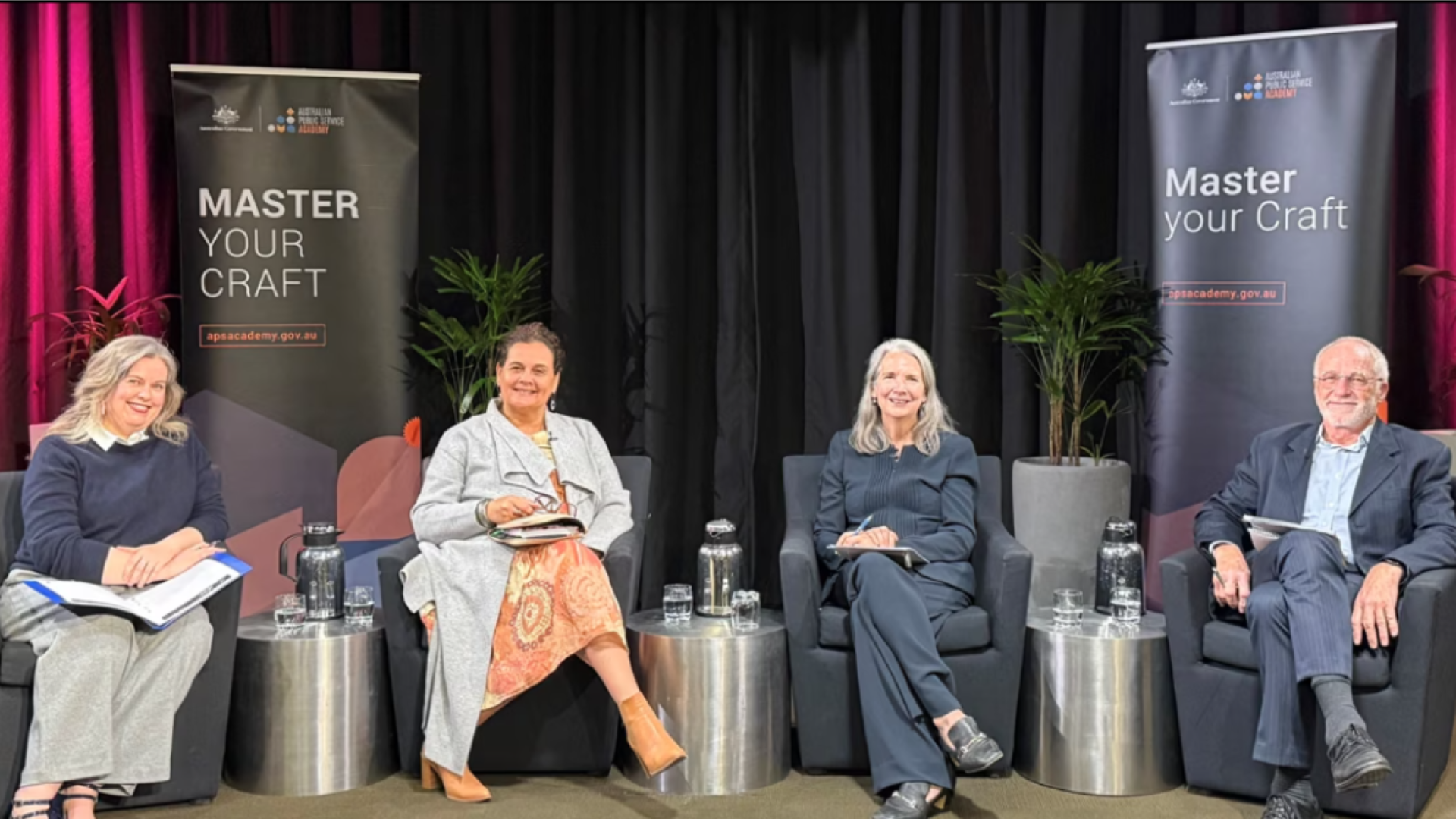 Photo of 4 panel members seated together in bucket chairs on stage facing the camera inside an auditorium. 