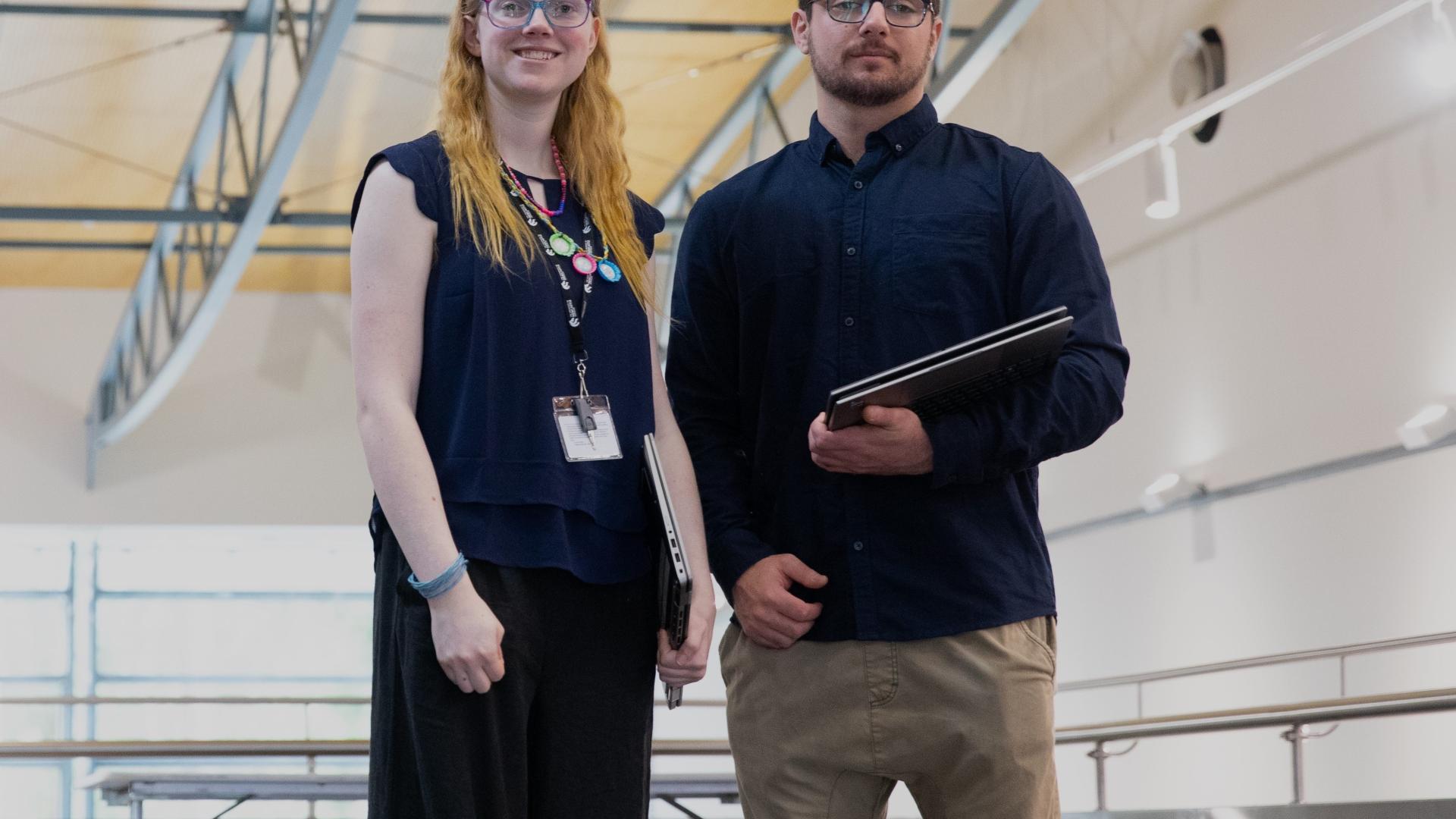 An image of 2 APS Academy Campuses graduates – woman and man - standing facing the camera inside a large auditorium. 