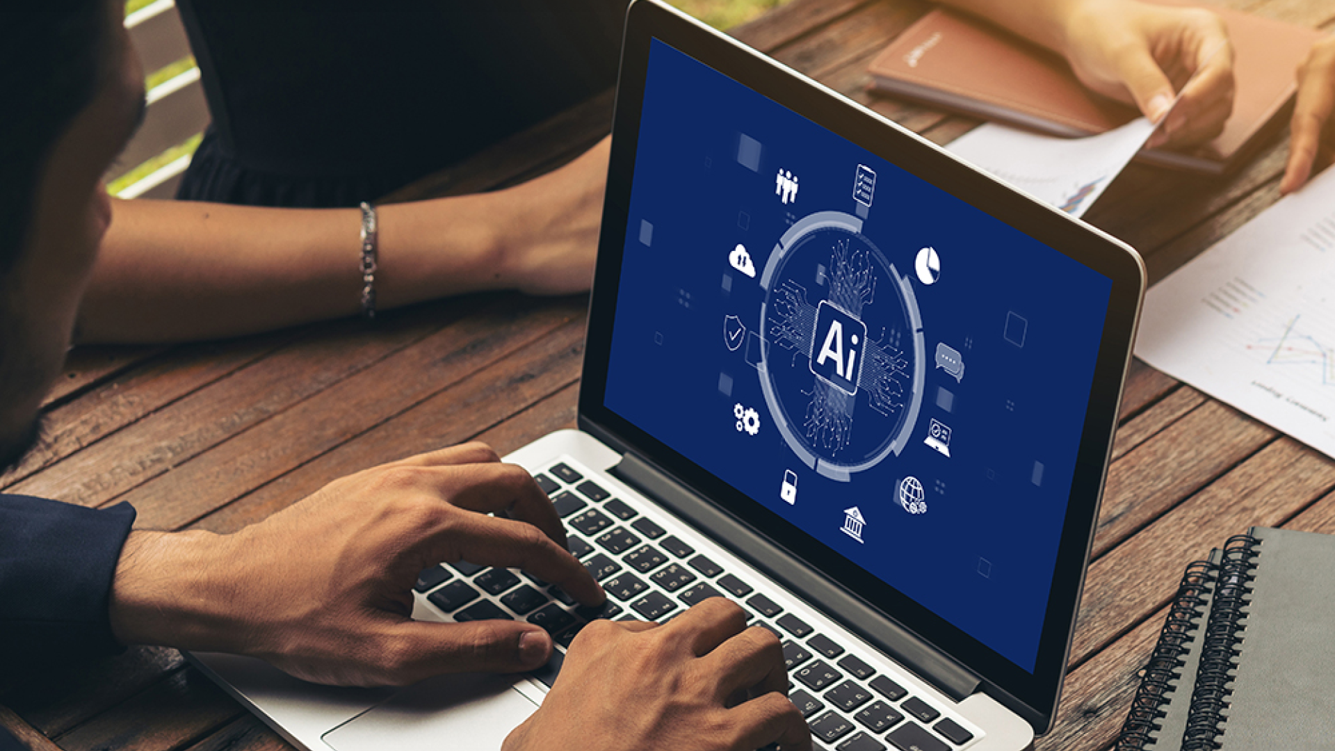 A person typing on a laptop displaying an artificial intelligence (AI) graphic, while others review documents at a wooden table 