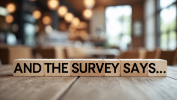 The image features 6 wooden blocks arranged horizontally on a wooden surface in a brightly lit room. Each block displays a word, collectively forming the phrase ‘AND THE SURVEY SAYS...’. The background is blurred with warm lighting, directing viewers’ attention towards the text. 