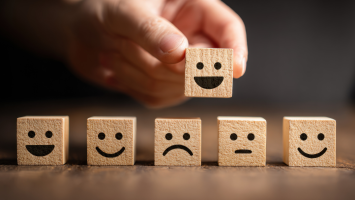The image features 6 small wooden cubes. Each cube has a simple, black emoticon face. The cubes are arranged in a row on a wooden surface with a dark background. A hand is picking up the fourth cube with a smiling face. 