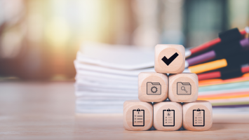 The image shows 6 wooden blocks stacked in a pyramid on a wooden surface. Each block has a black icon. The top block displays a check mark. The middle row contains 2 blocks with folder icons. The bottom row has 3 blocks with clipboard icons. The background is warm and has a stack of documents with colorful tabs. 
