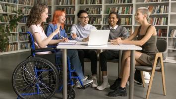 Group of people sitting around a table, in discussion, in what looks like a library or study space)