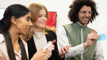 Three young adults stand together smiling and engaging in a collaborative activity. One person holds a brown marker, another holds a red marker, and the third holds a blue sticky note and an orange marker. They appear to be brainstorming or participating in a workshop in a bright indoor setting.