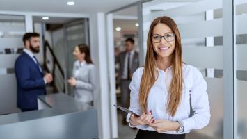 An image of an Executive Assistant standing in the foreground in an office setting with 2 other people in the background. 