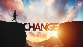 A silhouette of a person walks from one cliff edge to another using the word "CHANGE" as a bridge, set against a dramatic sunrise or sunset sky.