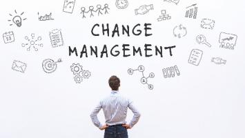 A person stands with hands on hips looking at the words "CHANGE MANAGEMENT" written on a white wall, surrounded by various hand-drawn business and planning icons.