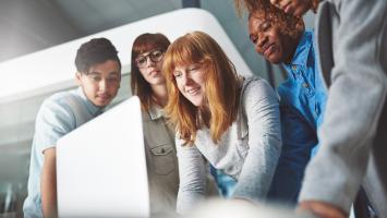 An image of 6 people gathered around a table looking at the laptop screen on it.