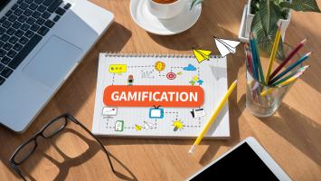 Notebook labelled "Gamification" on a desk with a laptop, coffee, stationery and illustrated icons representing learning, engagement and digital concepts.