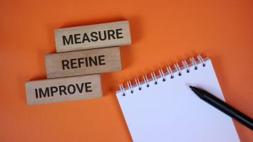 orange background with wooden blocks reading measure, refine, and improve beside a spiral notebook and black pen, symbolizing strategic planning, workflow optimization, and ongoing improvement. 